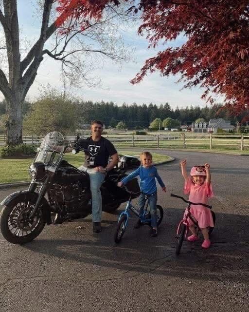 A man rides a flame-painted Harley down a winding road, his two children holding tight behind him, the sunset casting a golden glow over the scene.