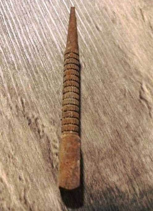 A close-up of a nail punch being used to drive a nail head below the surface of a wooden board, with a hammer poised to strike the opposite end.