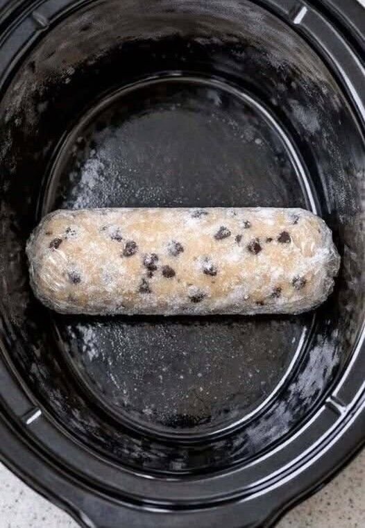A slow cooker filled with a warm, gooey cookie cobbler, topped with melted white chocolate chips and fruit pie filling, served in a bowl with a spoon.