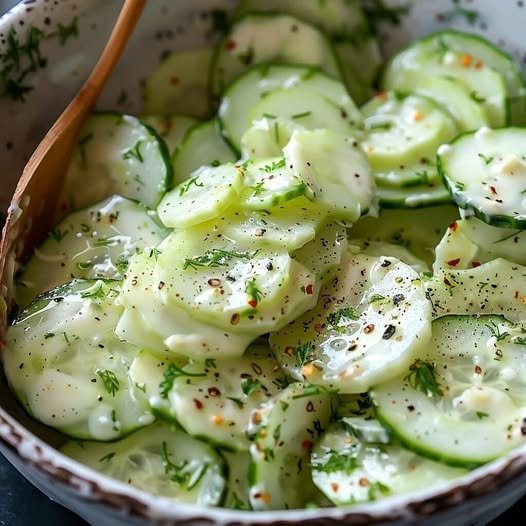 A bowl of creamy cucumber salad garnished with fresh dill, surrounded by sliced cucumbers, red onions, and a wooden spoon, set on a rustic table with a checkered cloth.