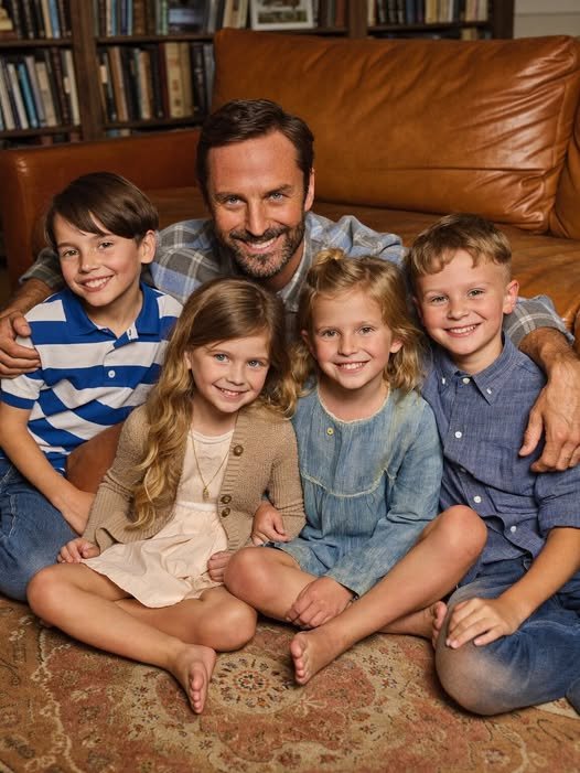 A man kneels beside four children, all smiling as they hold a framed photo of their late parents, their expressions filled with love and hope.