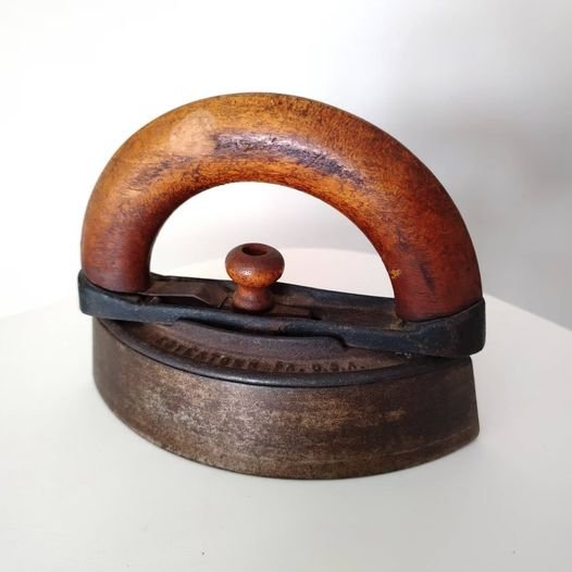 A vintage charcoal iron with its lid open, revealing the chamber for glowing embers, resting on an old wooden table beside a piece of fabric.