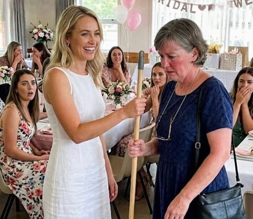 Future mother-in-law holding a mop at a bridal shower while guests react in shock after a surprising gift reveal.